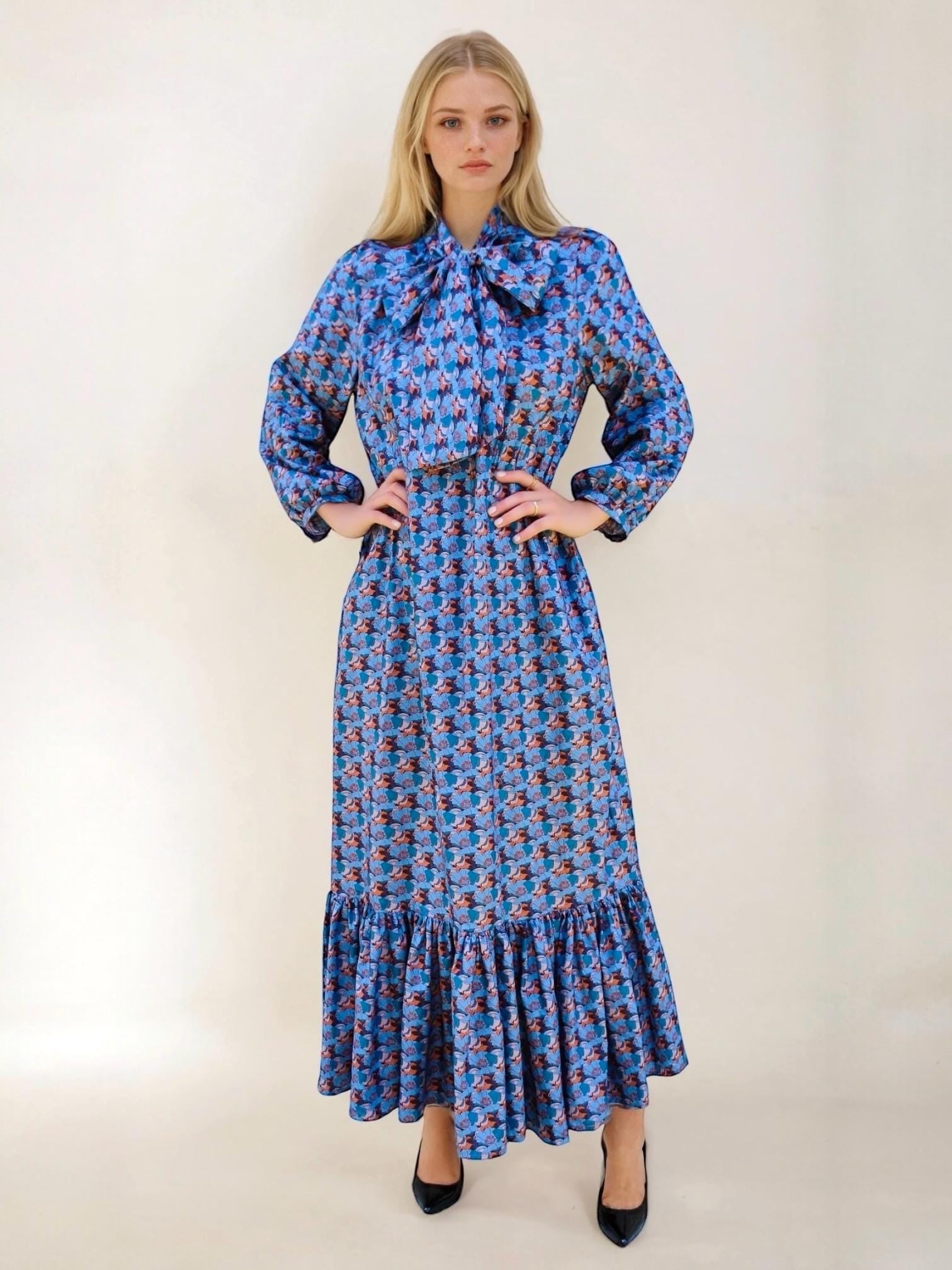 May include: A long, blue dress with a floral pattern. The dress features long sleeves, a large bow at the neckline, and a ruffled hem. The dress is worn with black heels.