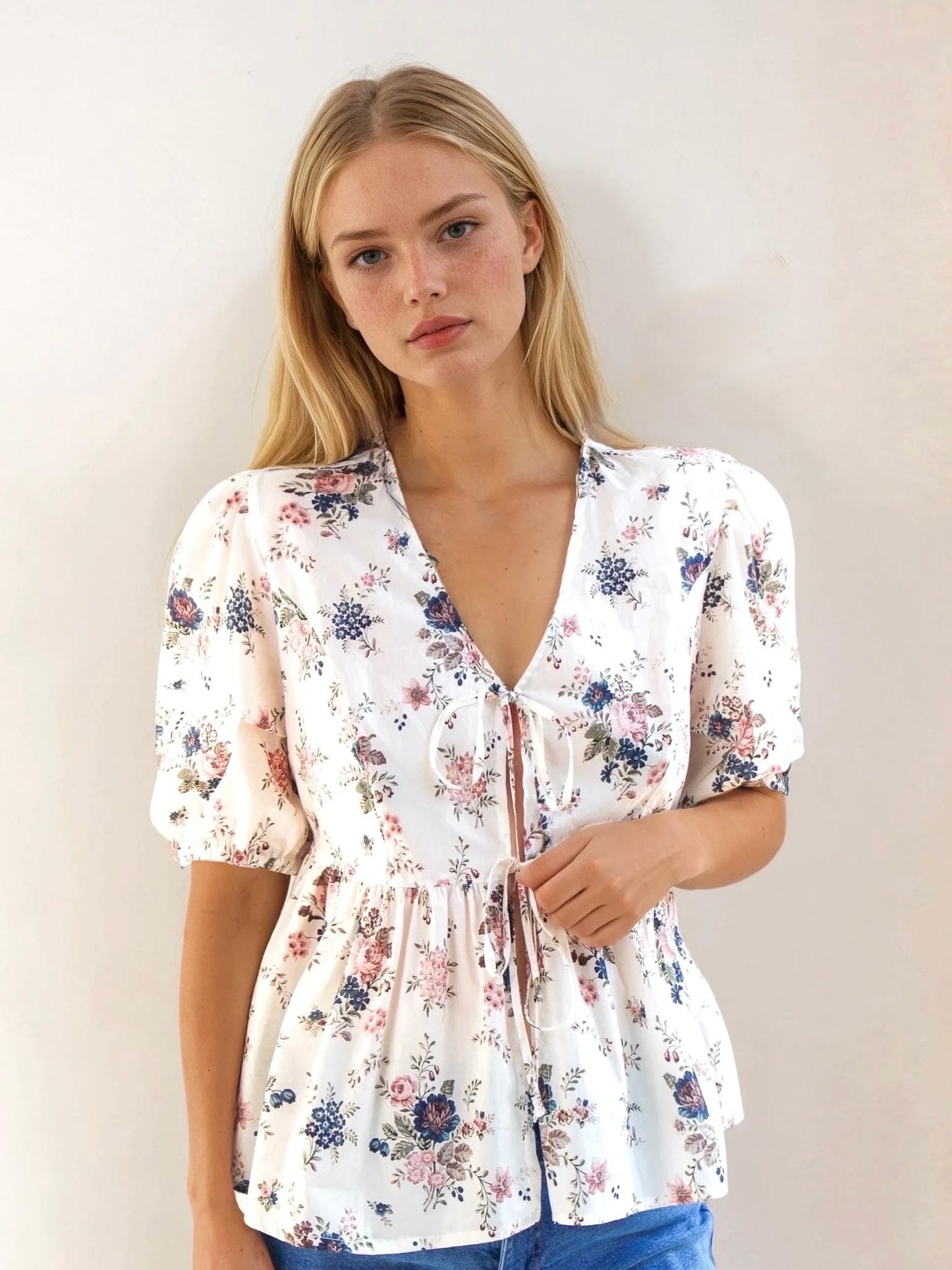 May include: A white floral print blouse with a V-neckline and short, puffed sleeves. The blouse features a tie closure at the bust and is adorned with pink, blue, and purple floral patterns. The fabric appears to be lightweight.