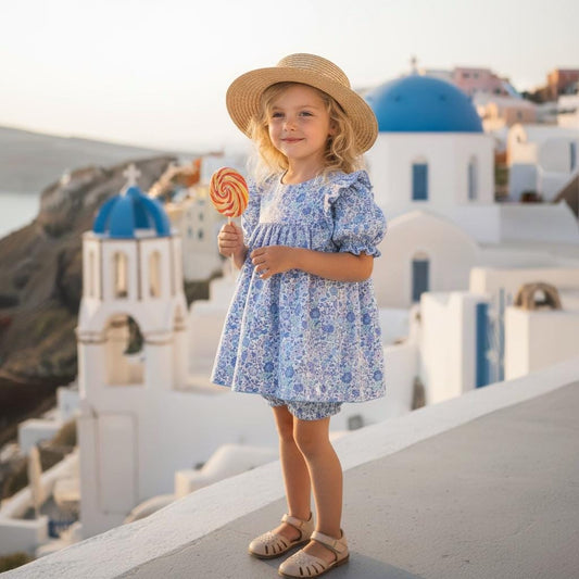 May include: A young child wearing a blue floral dress, matching bloomers, and a straw hat, holding a swirl lollipop. The outfit is paired with beige sandals. The background features white buildings with blue domes.