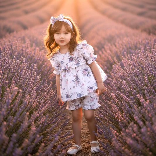 May include: A young child wearing a white floral print outfit, including a top with ruffled sleeves and shorts. The outfit is paired with a matching headband and white shoes. The child is standing in a field of lavender.