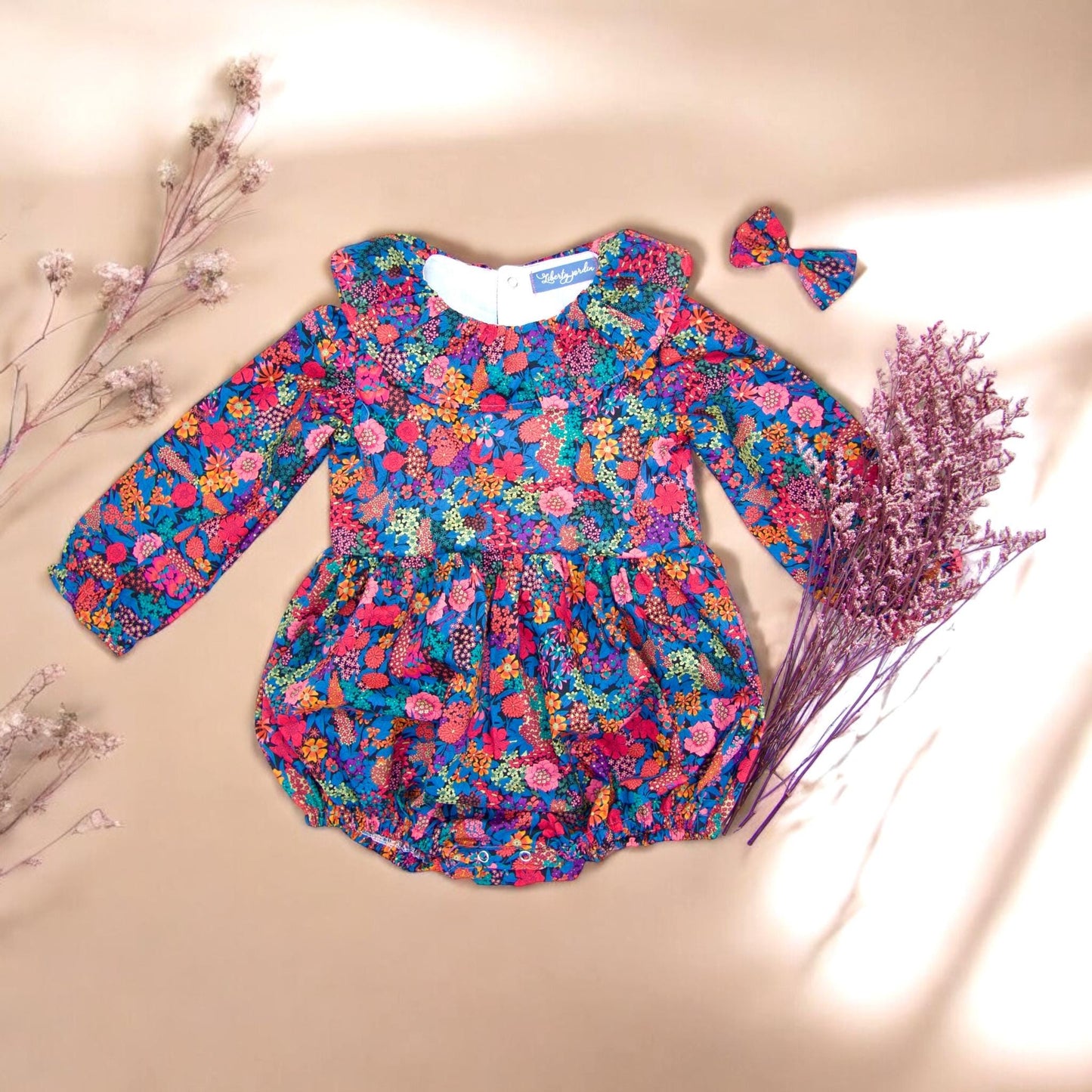 May include: A blue and pink floral print baby onesie with a white ruffled collar and long sleeves. The onesie has a bow headband.