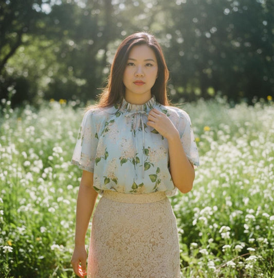 May include: A woman wearing a light blue floral top with a high neckline and a beige lace skirt stands in a field of white flowers. The top has short sleeves and a delicate floral pattern. The skirt is knee-length.