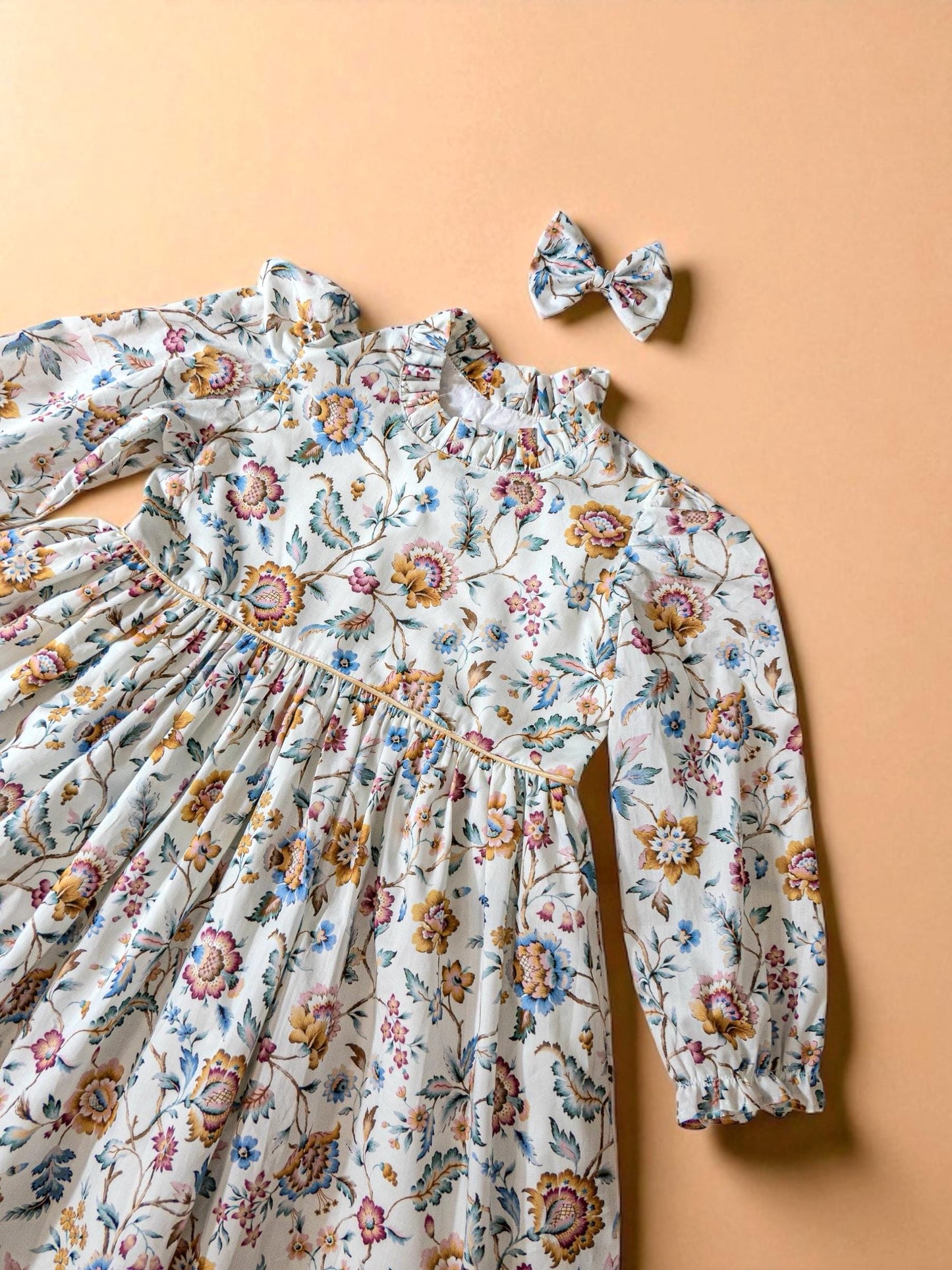 May include: A white floral print dress with long sleeves and a matching bow. The dress features a high ruffled collar and a gathered waist. The floral pattern includes blue, yellow, and pink flowers on a white background. The bow is the same floral pattern.