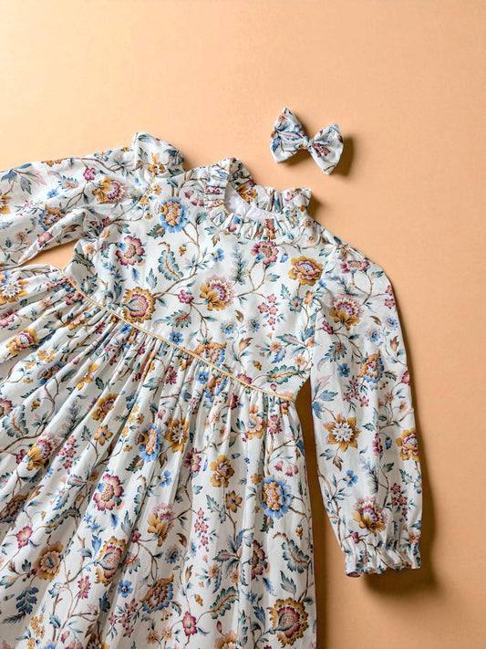 May include: A white floral print dress with long sleeves and a matching bow. The dress features a high ruffled collar and a gathered waist. The floral pattern includes blue, yellow, and pink flowers on a white background. The bow is the same floral pattern.
